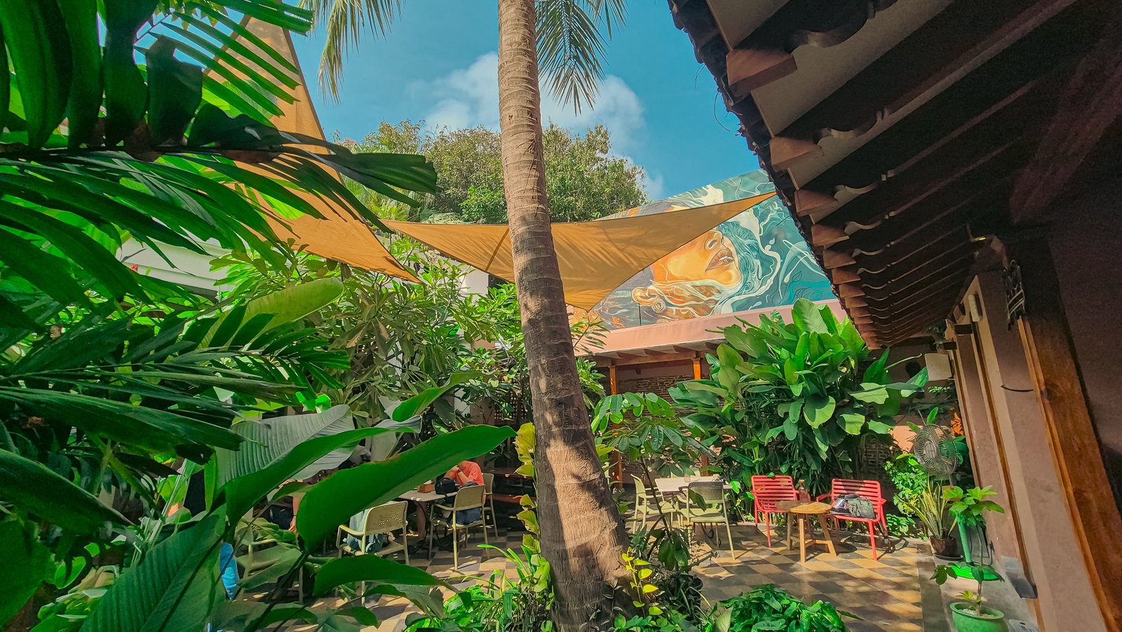 Cherry Pond, a lush biophilic courtyard in Puducherry with shade sails, a tall palm, tropical planting and casual seating